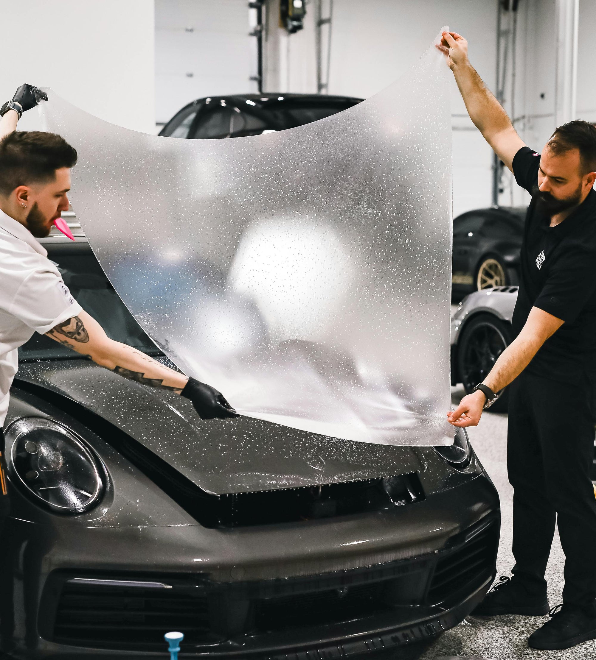 Two men working on a car in a garage, with one applying a protective film to the car's hood.